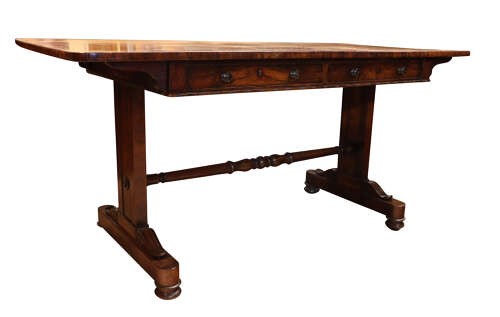 A William IV rosewood library table featuring a rectangular top, two frieze drawers with turned wooden knobs, trestle end supports, and a central turned stretcher. View IV