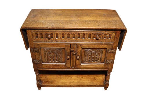 A warm brown carved oak side cabinet featuring drop-leaf top extensions, arcaded carved cupboard doors, turned legs, and a lower pot board shelf. View II