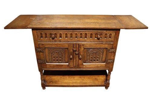 A warm brown carved oak side cabinet featuring drop-leaf top extensions, arcaded carved cupboard doors, turned legs, and a lower pot board shelf. View III