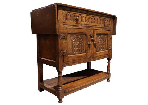 A warm brown carved oak side cabinet featuring drop-leaf top extensions, arcaded carved cupboard doors, turned legs, and a lower pot board shelf. View IV