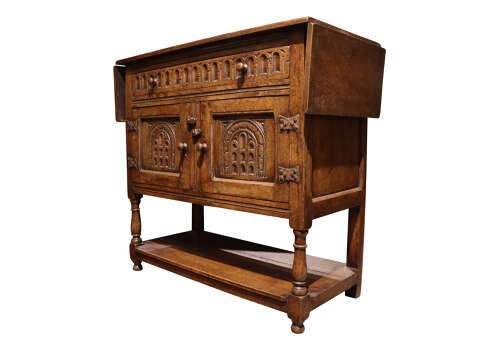 A warm brown carved oak side cabinet featuring drop-leaf top extensions, arcaded carved cupboard doors, turned legs, and a lower pot board shelf. View V