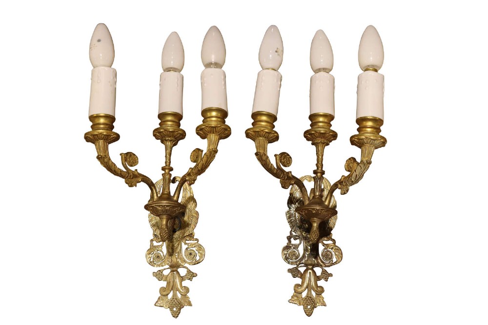 A pair of early 20th-century three-branch cast brass wall sconces featuring ornate acanthus leaf scrollwork and candle-style bulb fittings.