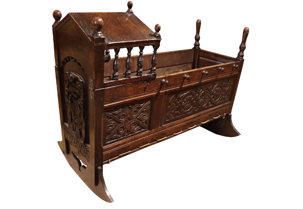 A dark carved oak rocking cradle featuring a hooded top with turned spindles, carved relief side panels, and curved rockers at the base.