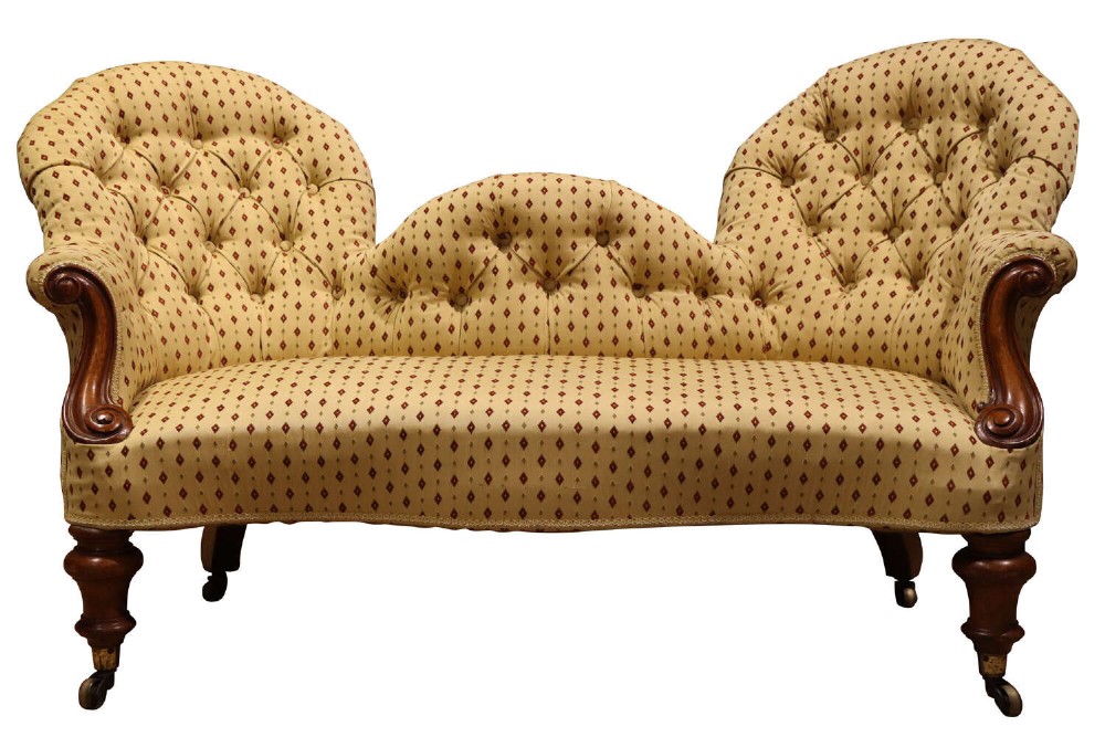 A Victorian mahogany double-ended settee featuring deep-buttoned patterned yellow upholstery, carved scroll arms, and turned legs with brass castors.