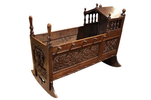 A dark carved oak rocking cradle featuring a hooded top with turned spindles, carved relief side panels, and curved rockers at the base. View IV