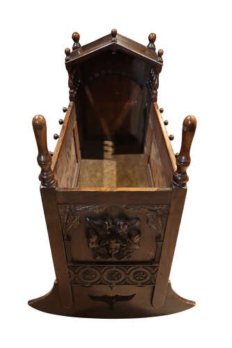 A dark carved oak rocking cradle featuring a hooded top with turned spindles, carved relief side panels, and curved rockers at the base. View VI