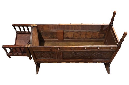 A dark carved oak rocking cradle featuring a hooded top with turned spindles, carved relief side panels, and curved rockers at the base. View VII