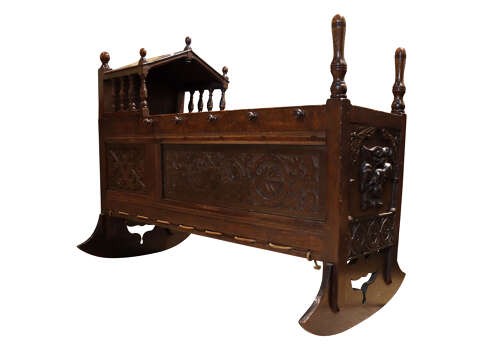 A dark carved oak rocking cradle featuring a hooded top with turned spindles, carved relief side panels, and curved rockers at the base. View VIII