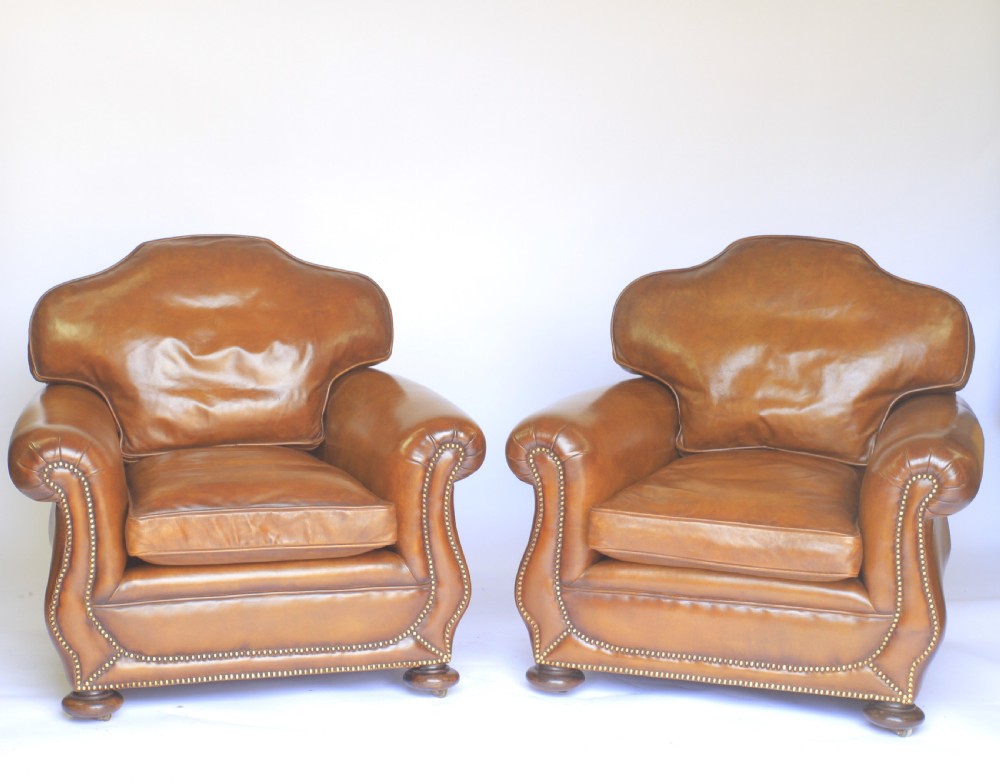 A pair of 1920s tan leather club armchairs featuring rolled arms, brass stud trimming, shaped crest backs, and turned wooden feet on castors.