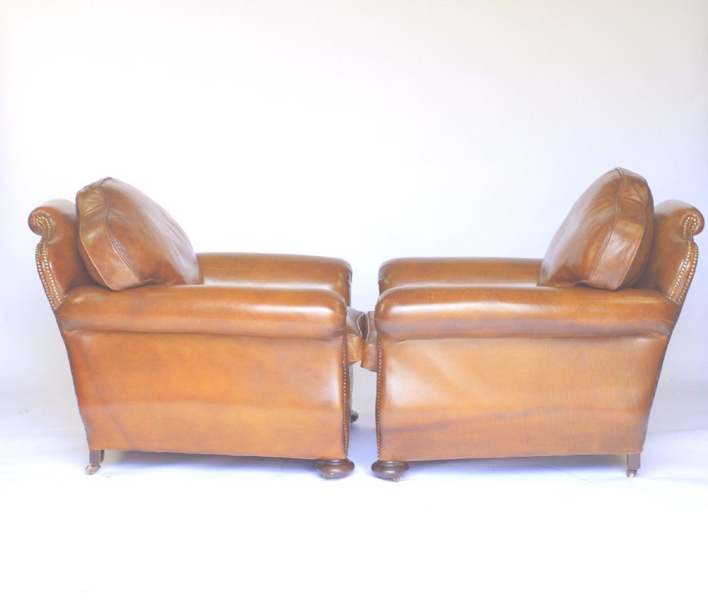 A pair of 1920s tan leather club armchairs featuring rolled arms, brass stud trimming, shaped crest backs, and turned wooden feet on castors. View V