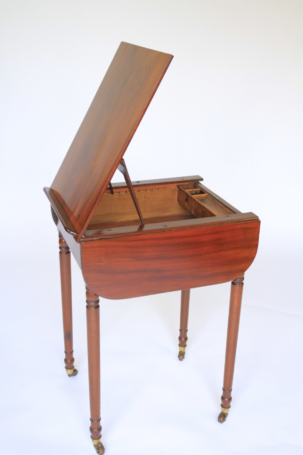 A Regency mahogany writing table with an adjustable ratchet top, side drop flap, internal fitted pen compartment, slim turned legs, and brass castors.