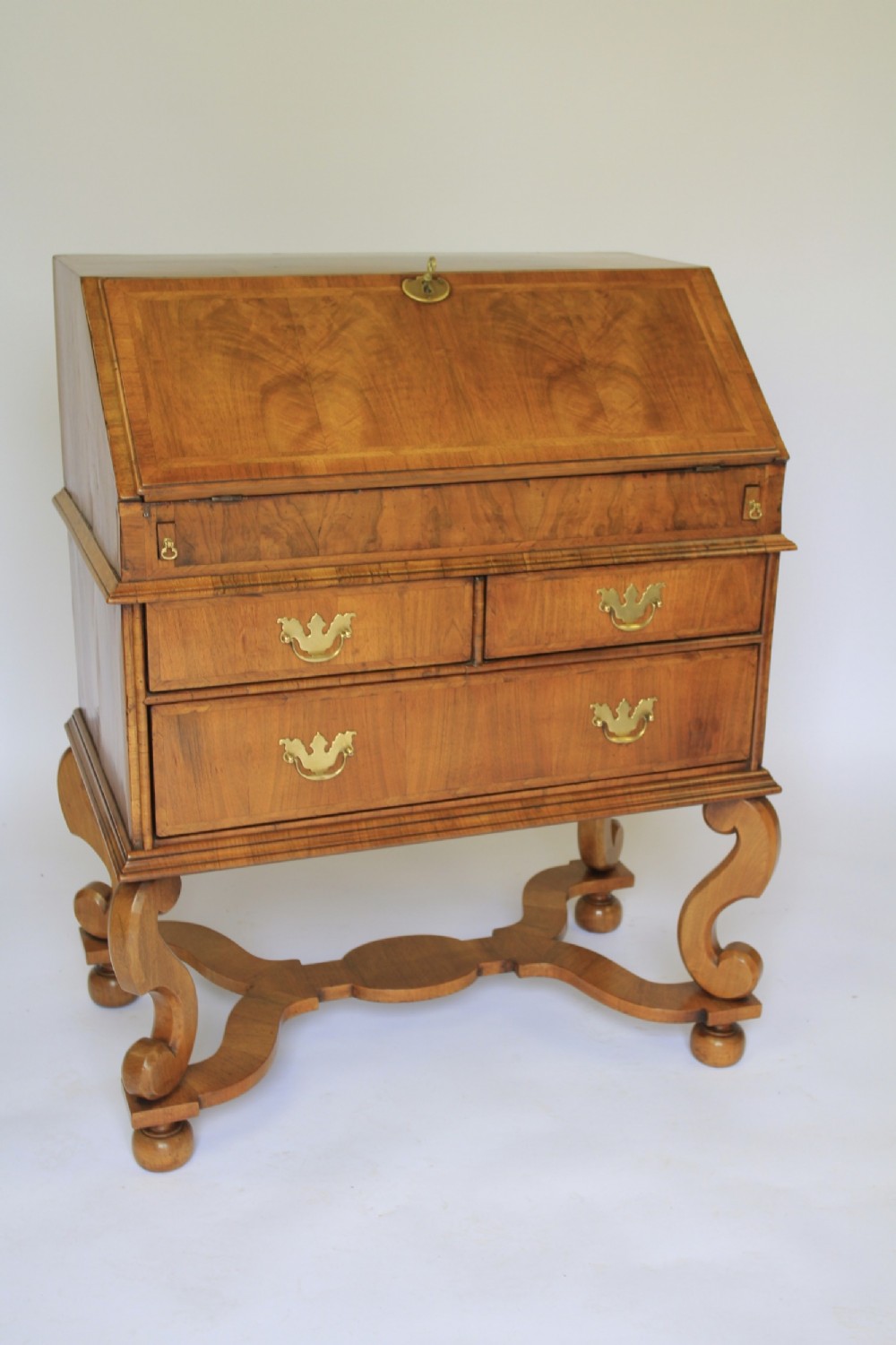 A William and Mary Revival walnut slant-front writing bureau with brass handles and carved scroll legs connected by a shaped stretcher.