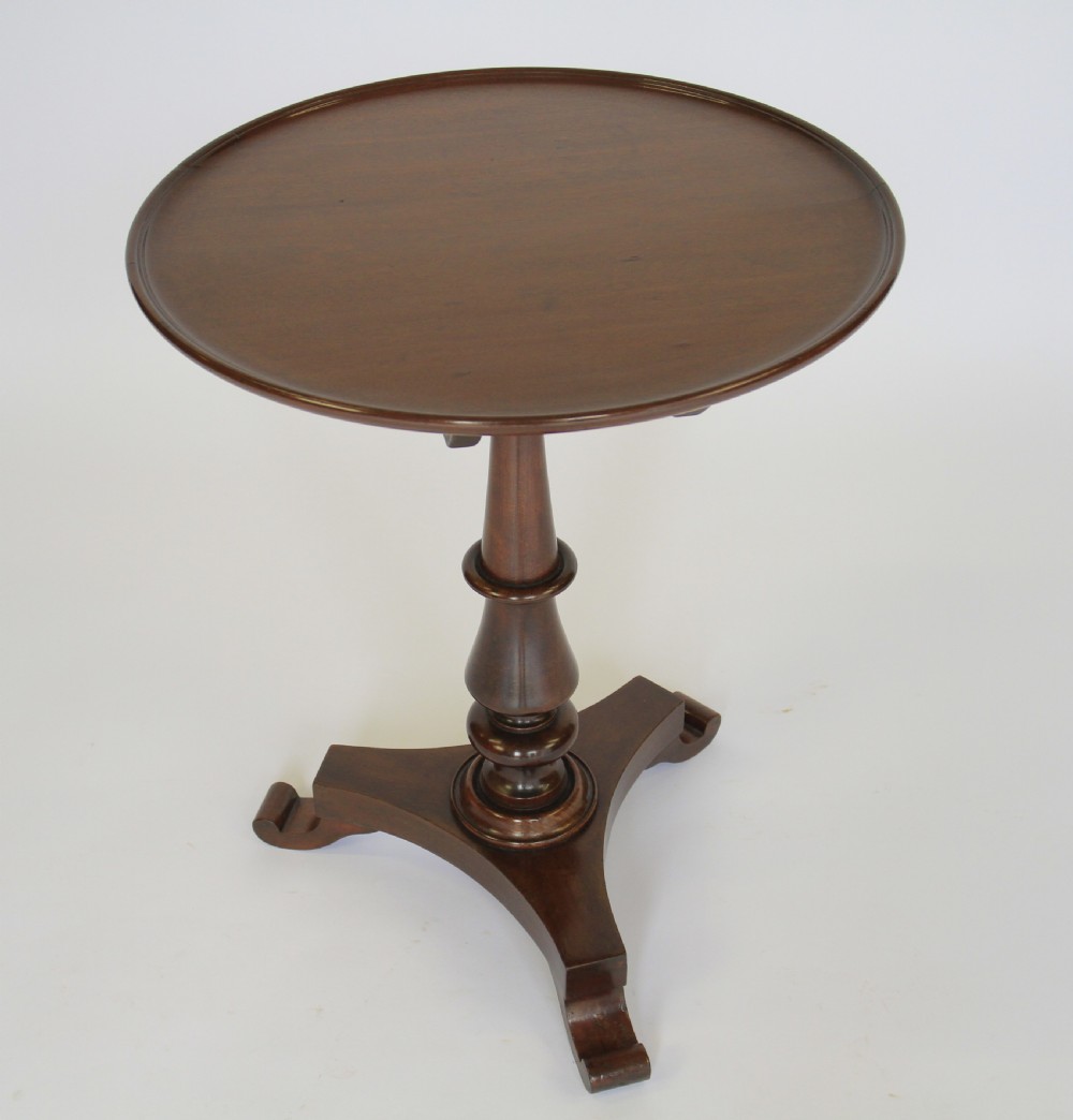 A William IV dark mahogany tilt-top table featuring a raised moulded edge rim, a turned central pedestal stem, and a tri-form platform base on scroll feet.