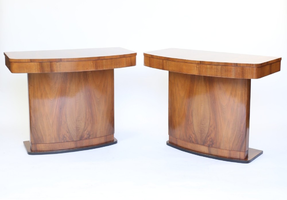 A pair of Art Deco console tables featuring curved walnut veneered tops, central pedestal bases with book-matched grain, and ebonised plinths.