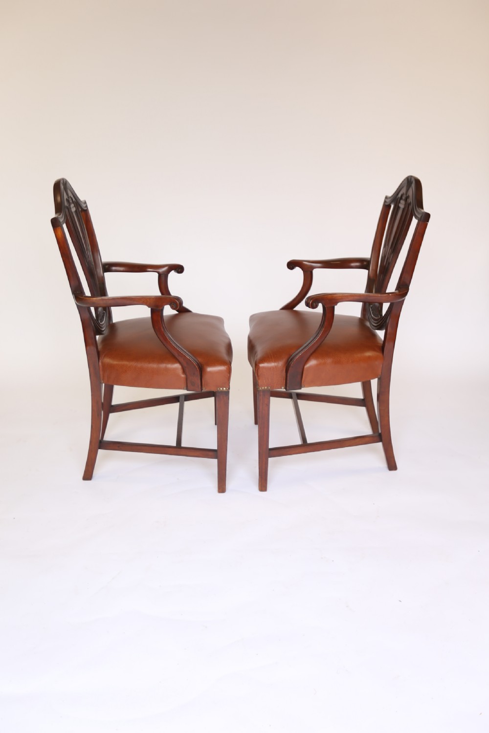 A pair of 1930s Hepplewhite style mahogany elbow chairs featuring carved shield backs and tan leather seats. View V
