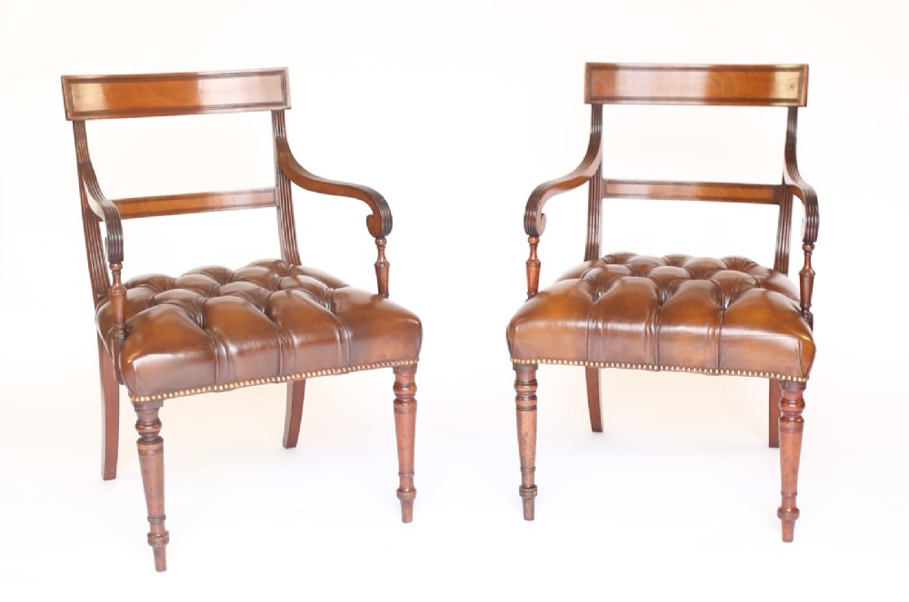 A pair of 19th-century mahogany elbow chairs featuring reeded arms, button-tufted brown leather seats with brass studding, and turned front legs.