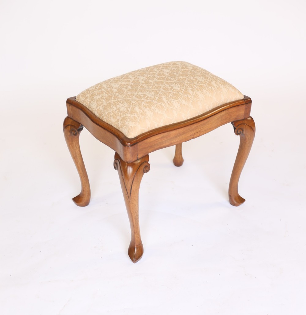 A 1930s Queen Anne style walnut dressing stool featuring carved cabriole legs and a cream patterned fabric seat.