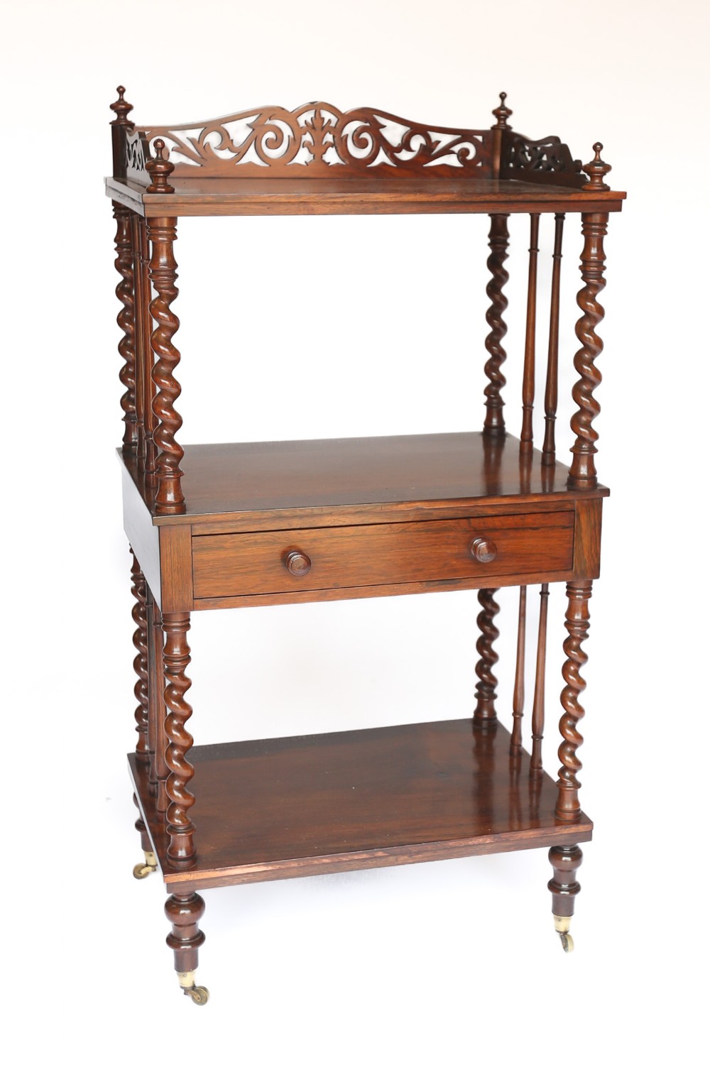A three-tier Victorian rosewood whatnot featuring barley twist corner supports, a pierced fretwork top gallery, a single central drawer, and brass castors.