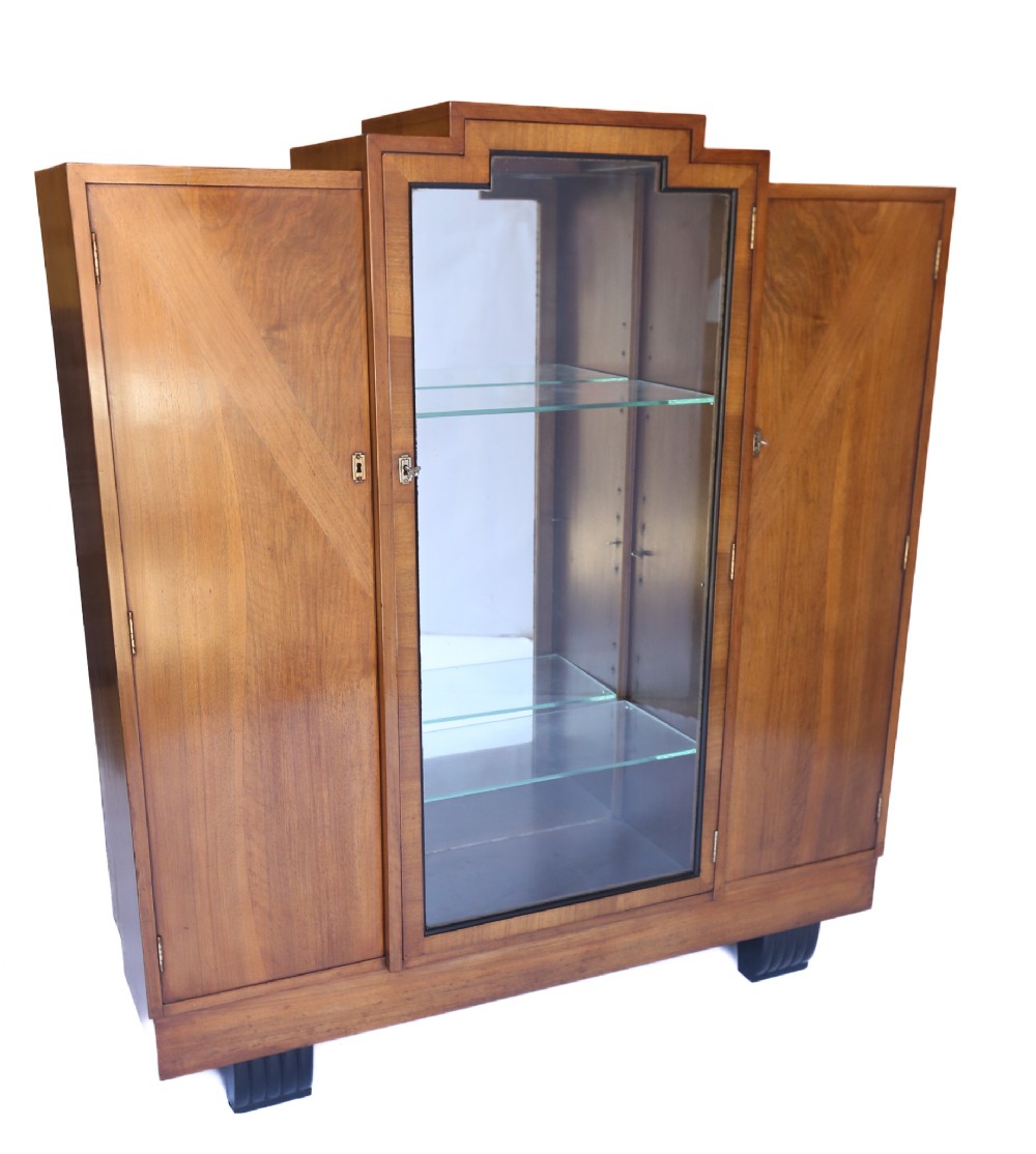 A 1930s Art Deco walnut display cabinet featuring a central glazed door with glass shelves, flanked by two veneered wooden cupboard doors and supported by ebonised feet.