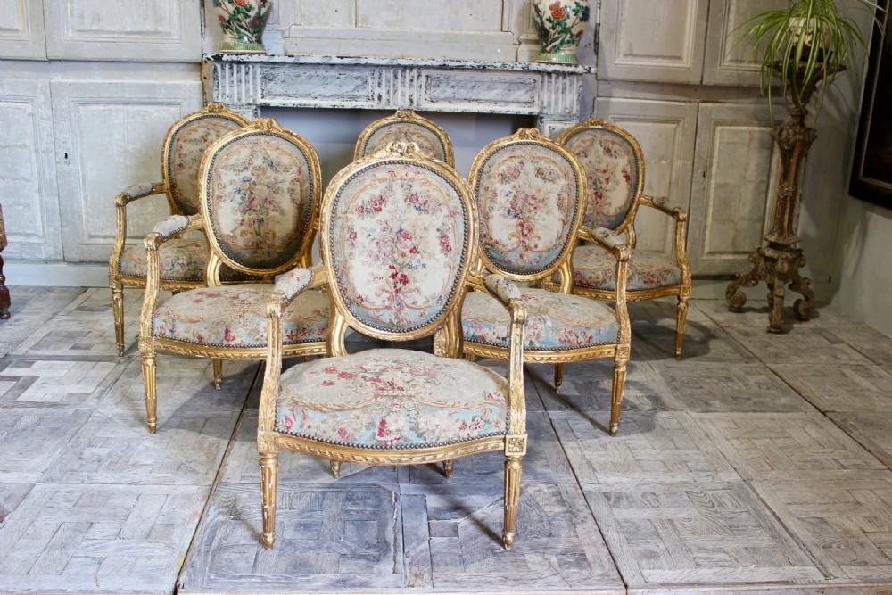 A set of six 18th-century Louis XVI French giltwood armchairs featuring oval medallion backs, carved armrests, fluted legs, and floral tapestry upholstery.