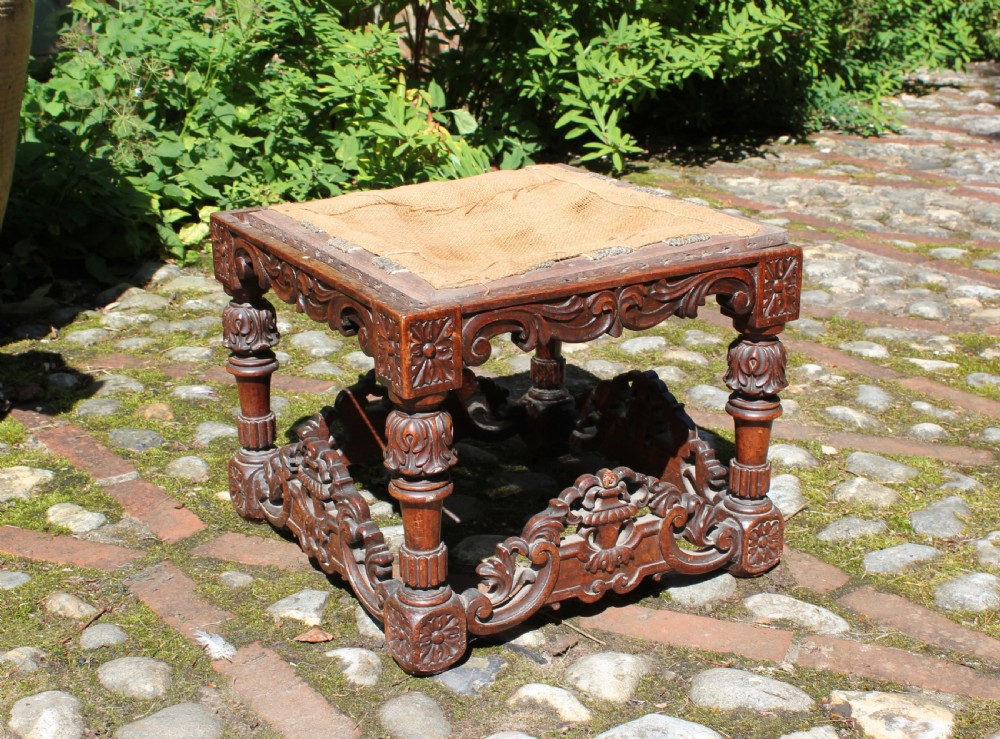 A square dark wood Batavian Baroque stool featuring carved floral aprons, turned baluster legs, pierced stretchers, and a hessian upholstered top.