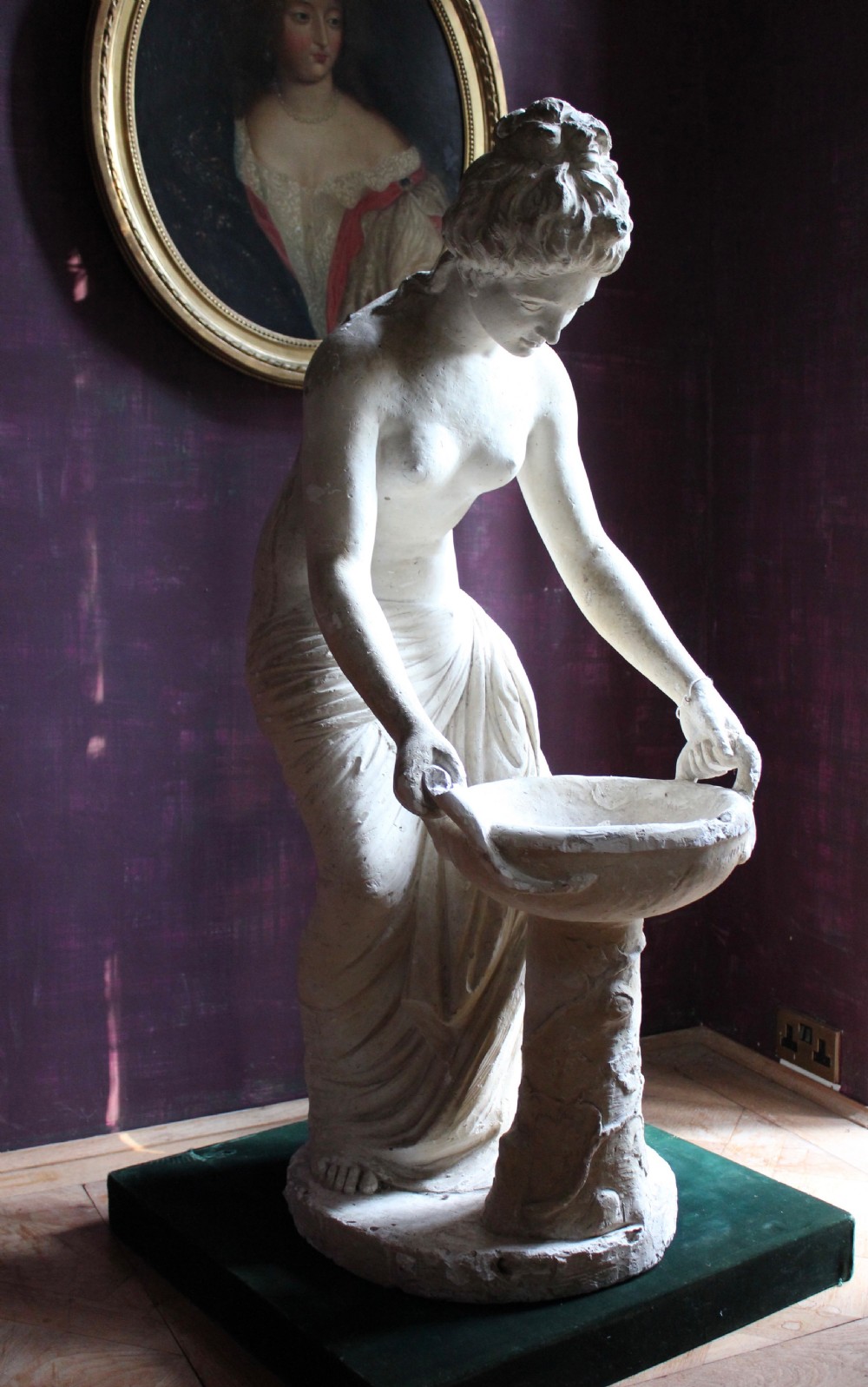 A full-length off-white plaster statue depicting a classical draped female figure standing beside a pedestal basin.