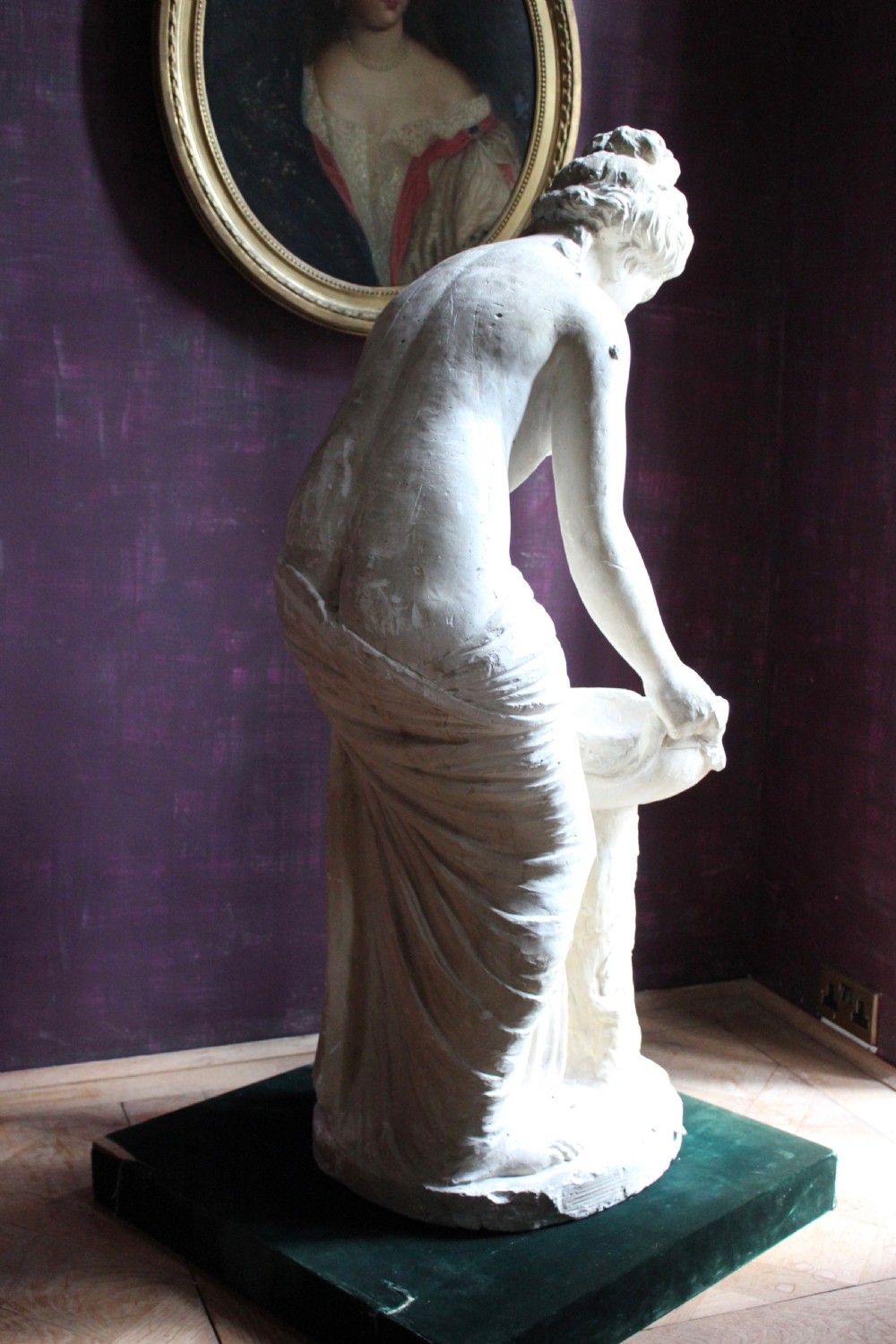 A full-length off-white plaster statue depicting a classical draped female figure standing beside a pedestal basin. View V