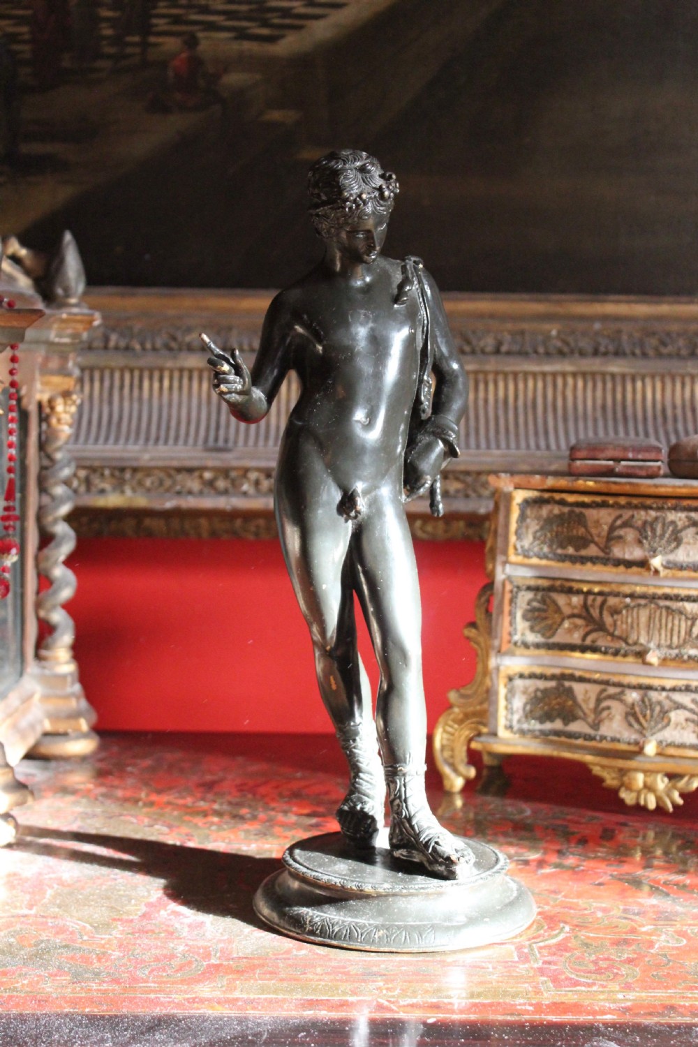 A dark patinated bronze statuette of Narcissus standing on a circular stepped base wearing laced sandals with drapery over his shoulder.