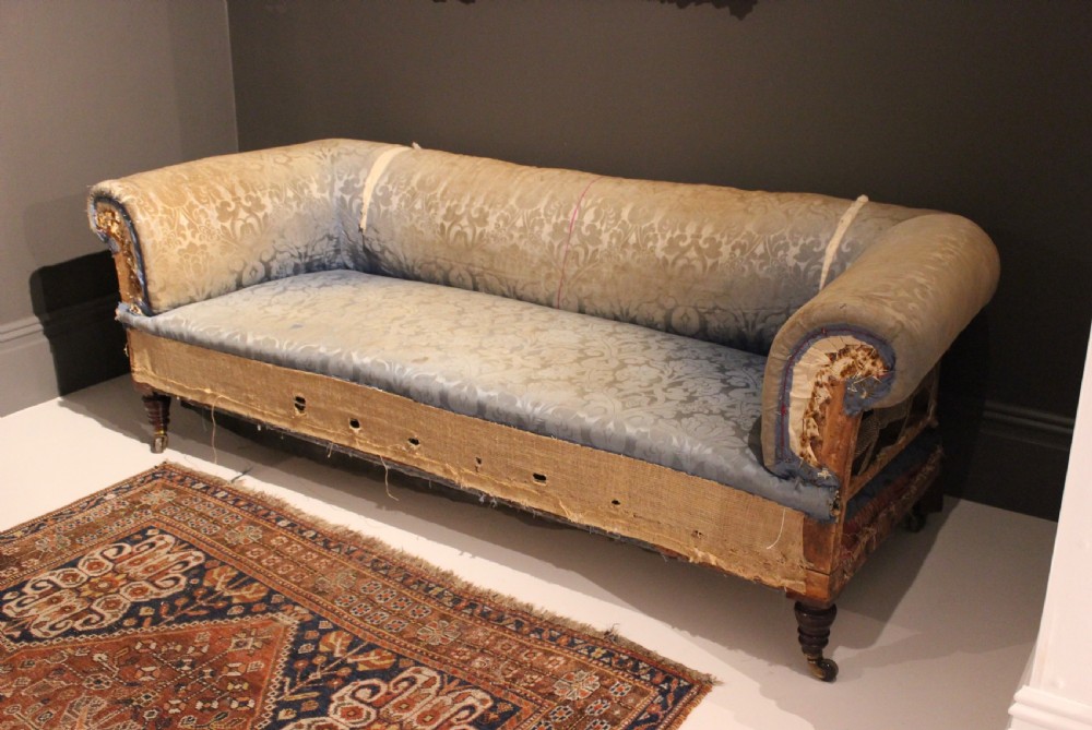 A Victorian Chesterfield sofa with rolled scroll arms, turned timber legs on brass castors, and patterned blue damask fabric over visible jute webbing. View IV