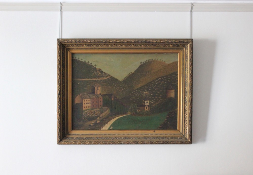 A 19th-century primitive oil painting depicting rolling hills, village buildings, and a winding path, housed in an ornate gilded frame.