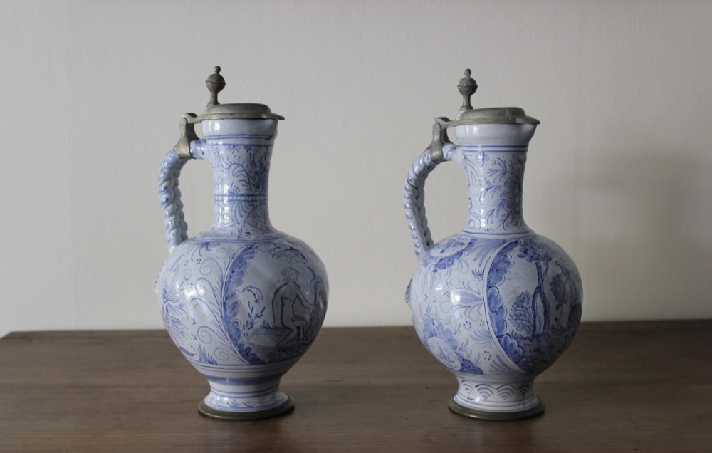 A matched pair of tin-glazed blue and white earthenware ewers featuring pewter hinged lids and twisted handles.