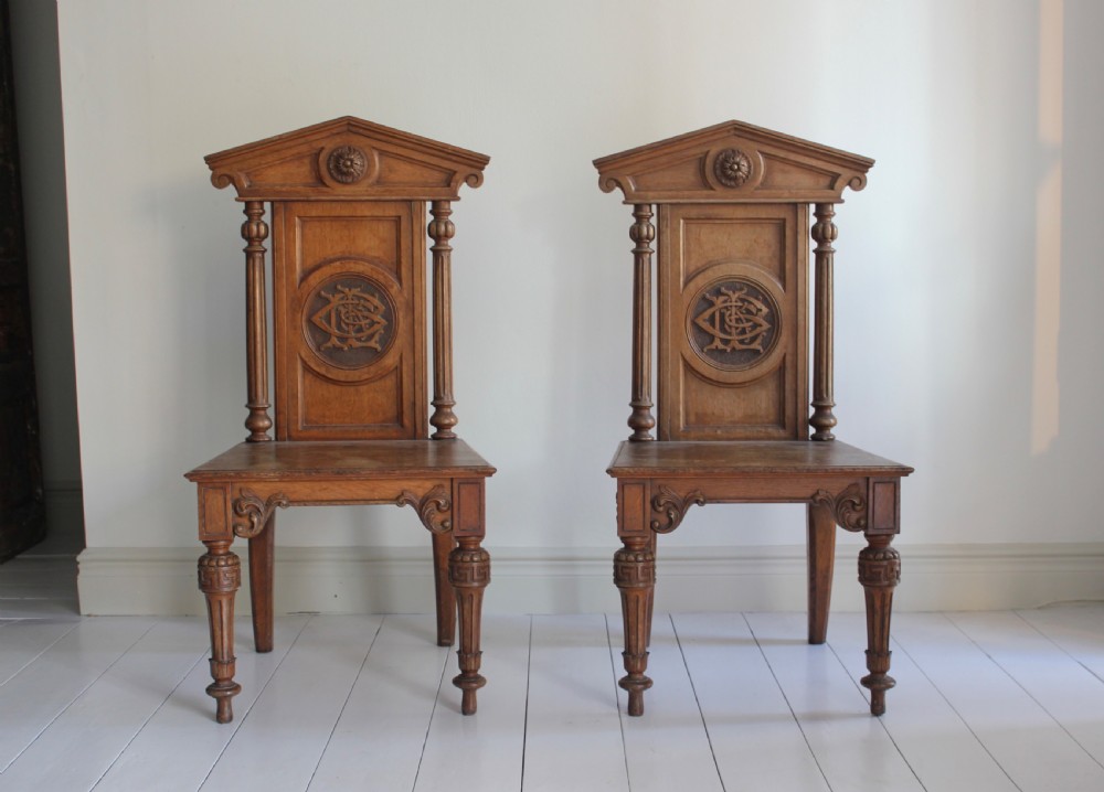 A pair of 19th-century English carved oak hall chairs featuring pediment backrests with central circular monograms and turned front legs. View II