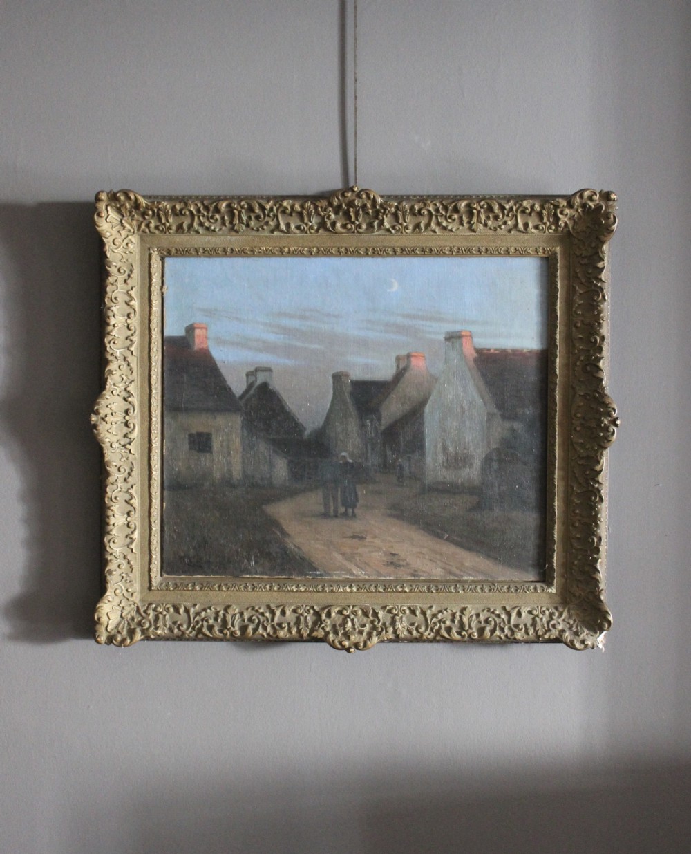 A 19th-century oil painting depicting a village street at twilight with cottages and a crescent moon, presented in an ornate giltwood frame.