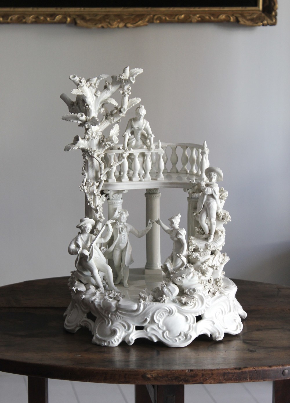 A white glazed porcelain sculpture depicting a multi-figure Rococo court scene with classical columns, a balcony, musicians, and a detailed foliate tree.