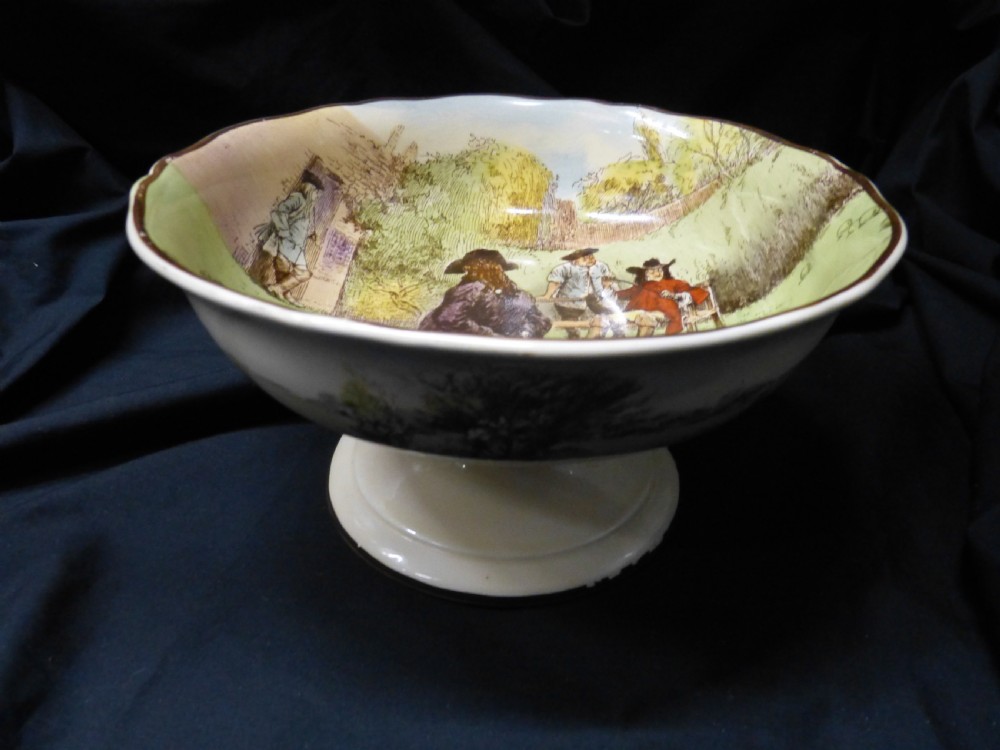 A footed ceramic bowl with a scalloped rim, featuring a printed pictorial scene of figures in a rural landscape on the interior surface.