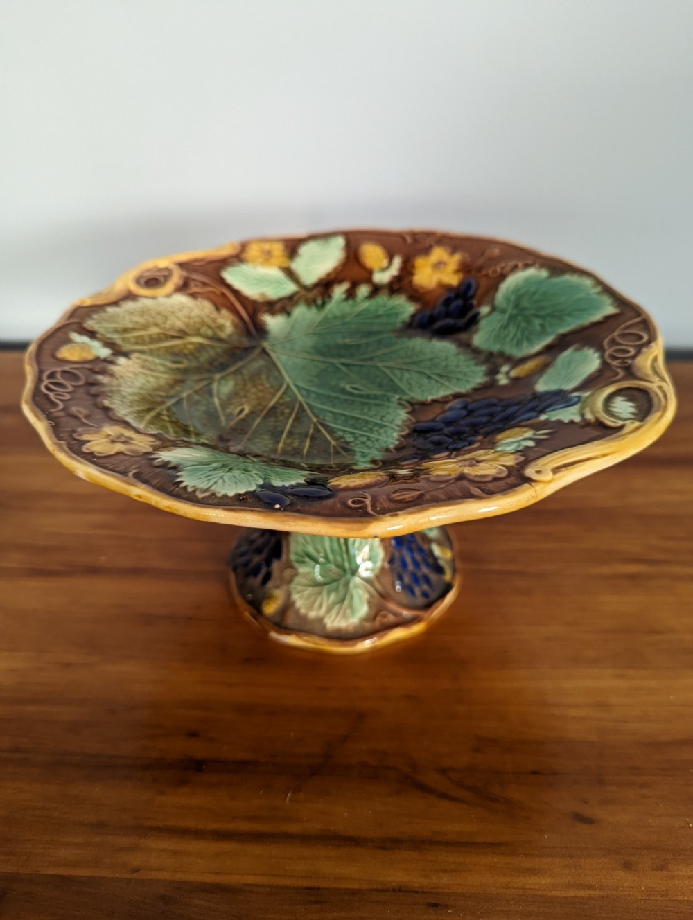 A Victorian majolica pedestal dish featuring moulded green leaves, blue berries, and yellow accents over a brown glazed earthenware surface.