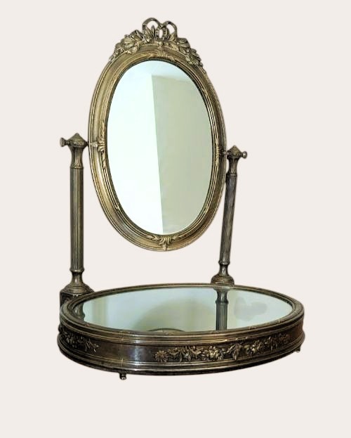 An antique French brass dressing mirror featuring a tilting oval frame with a ribbon crest, supported by fluted columns above a mirrored base.