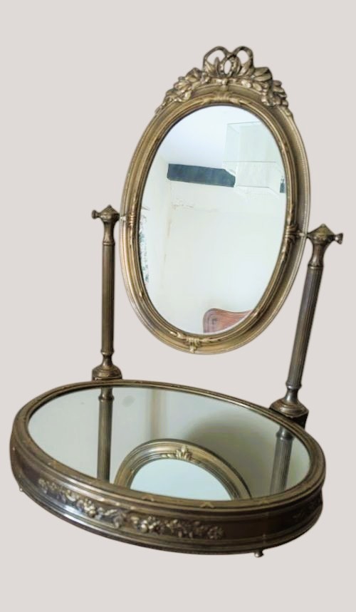 An antique French brass dressing mirror featuring a tilting oval frame with a ribbon crest, supported by fluted columns above a mirrored base. View III