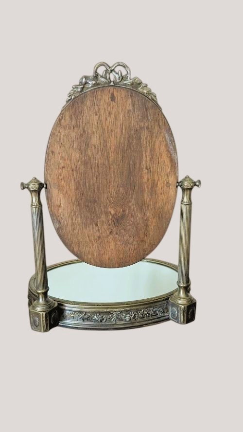 An antique French brass dressing mirror featuring a tilting oval frame with a ribbon crest, supported by fluted columns above a mirrored base. View IV