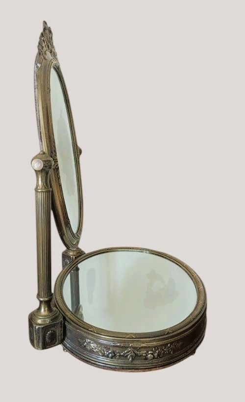 An antique French brass dressing mirror featuring a tilting oval frame with a ribbon crest, supported by fluted columns above a mirrored base. View VI