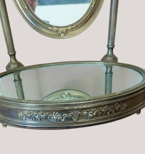 An antique French brass dressing mirror featuring a tilting oval frame with a ribbon crest, supported by fluted columns above a mirrored base. View VII