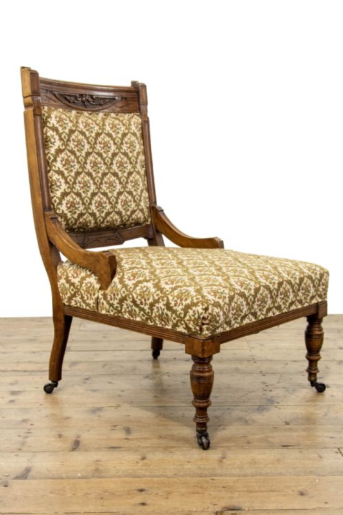 fig 1. An antique Edwardian low-slung chair featuring a carved timber frame, patterned textile upholstery, turned front legs, and castors.