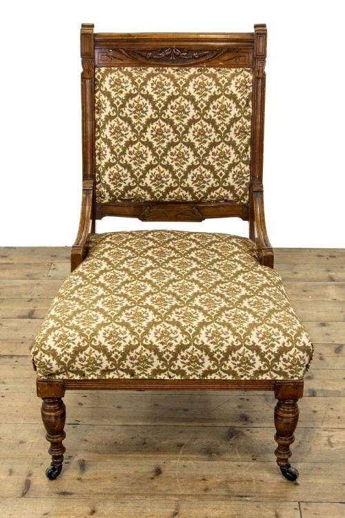 fig 2. An antique Edwardian low-slung chair featuring a carved timber frame, patterned textile upholstery, turned front legs, and castors.