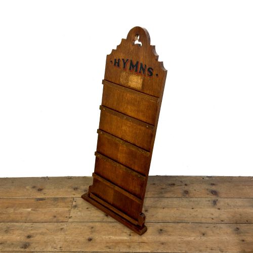 fig 1. An antique dark oak church hymn board featuring a trefoil cutout at the carved top, painted text reading HYMNS, multiple horizontal slots, and an accompanying wooden box.