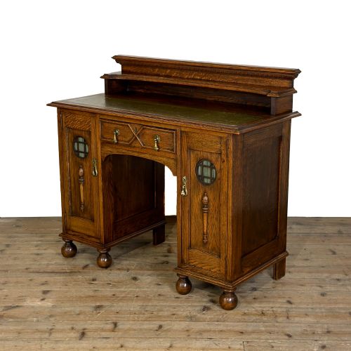 fig 1. An Arts and Crafts solid oak kneehole desk featuring a raised top gallery shelf, central drawer with geometric moulding, drop handles, and side cupboards with circular gridded glass panels.