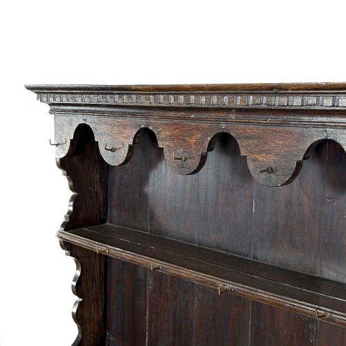 fig 1. A dark oak wall-mounted plate rack featuring three open shelves, a scalloped top frieze with hooks, and dentil moulding along the upper cornice.