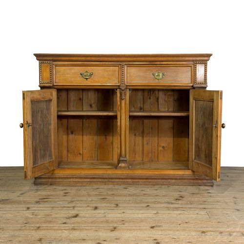 fig 1. An antique golden brown oak sideboard featuring two drawers with brass handles over two panelled cupboard doors with carved side pilasters.