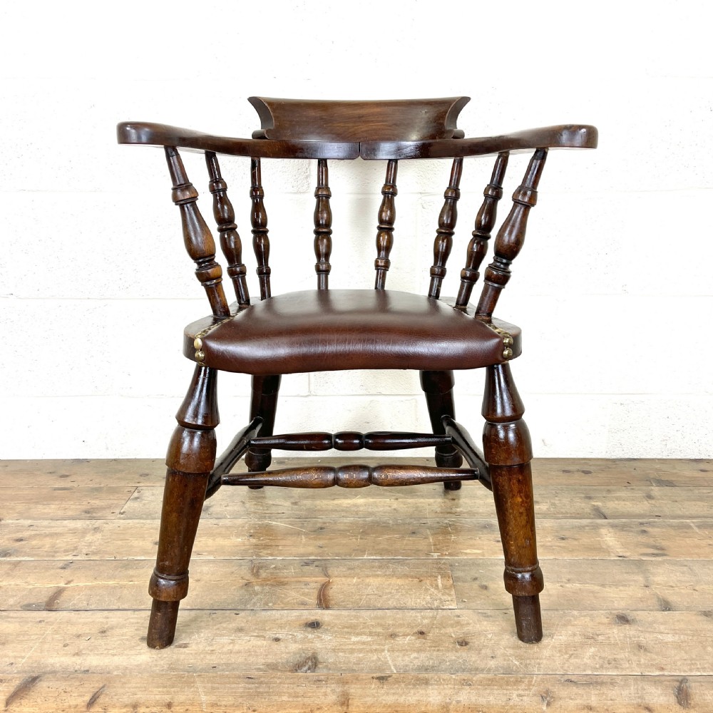An antique dark brown wooden smokers bow armchair featuring turned spindles, a curved top rail, and a brown leather upholstered seat secured with brass studs.