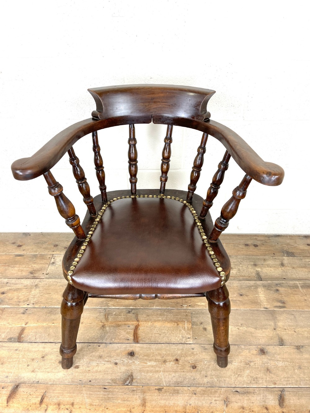 An antique dark brown wooden smokers bow armchair featuring turned spindles, a curved top rail, and a brown leather upholstered seat secured with brass studs. View III