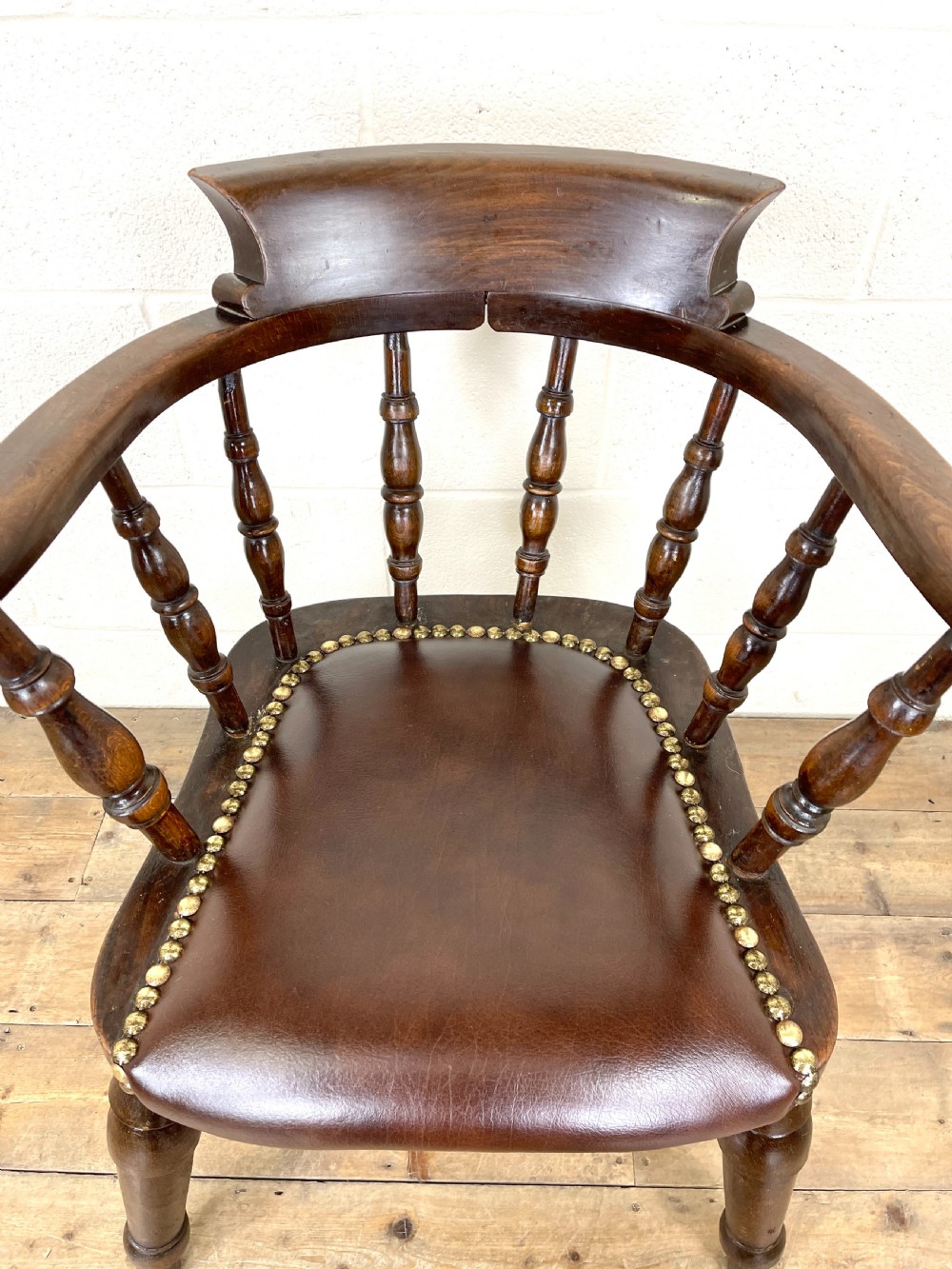 An antique dark brown wooden smokers bow armchair featuring turned spindles, a curved top rail, and a brown leather upholstered seat secured with brass studs. View V